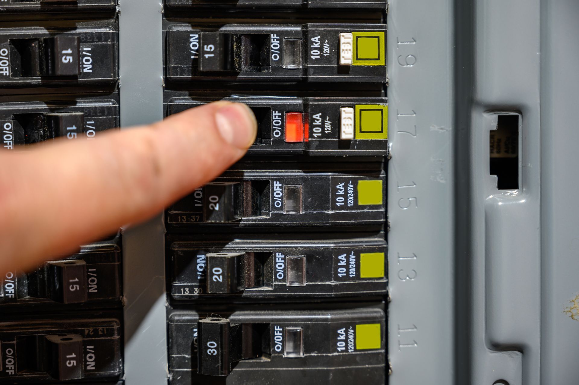 Finger pointing at a red breaker switch in an electrical panel with yellow toggles.