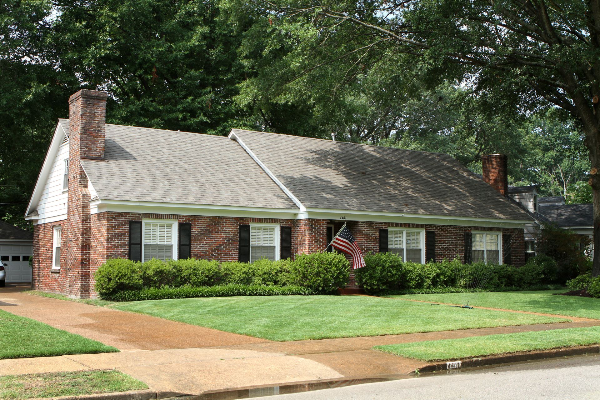 Single-story brick house with green lawn, shrubs, and trees along a quiet street Single-story brick house with green lawn, shrubs, and trees along a quiet street
