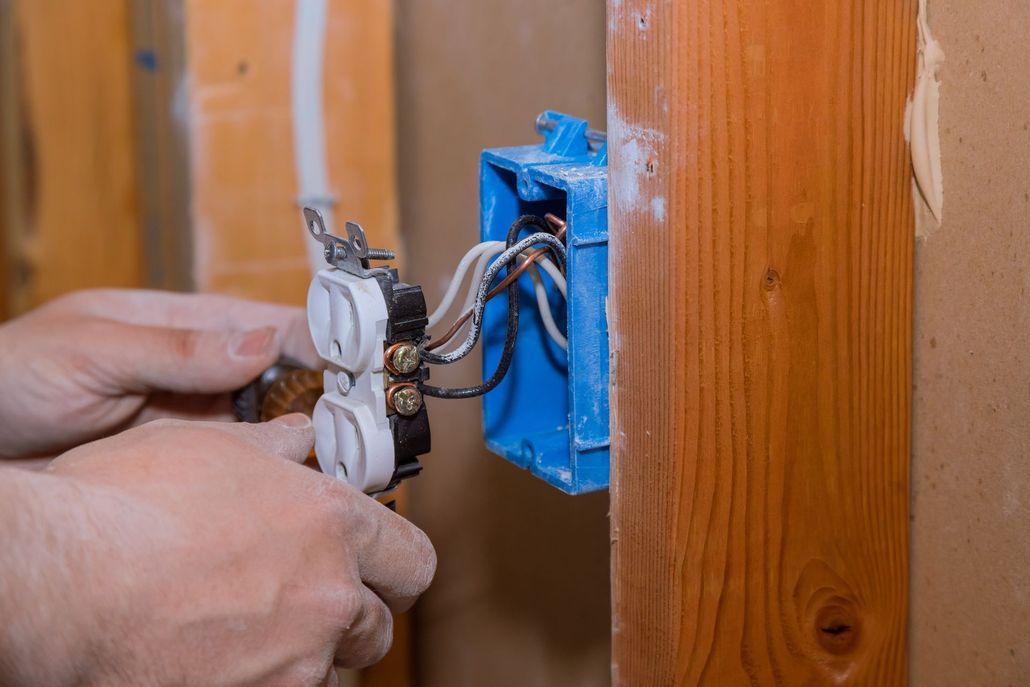 Hands wiring a white electrical outlet into a blue wall box beside wood paneling.