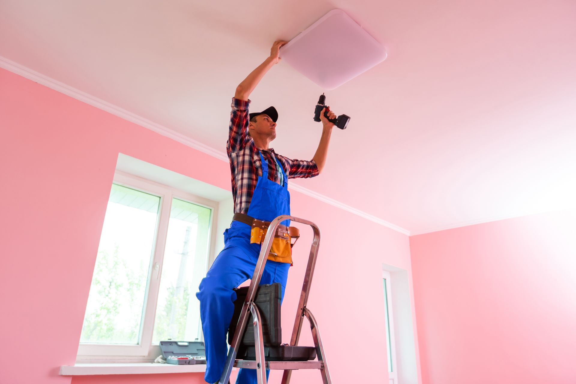 Worker on ladder painting a pink ceiling, holding a brush and paint tray in a room with white trim. Worker on ladder painting a pink ceiling, holding a brush and paint tray in a room with white trim.