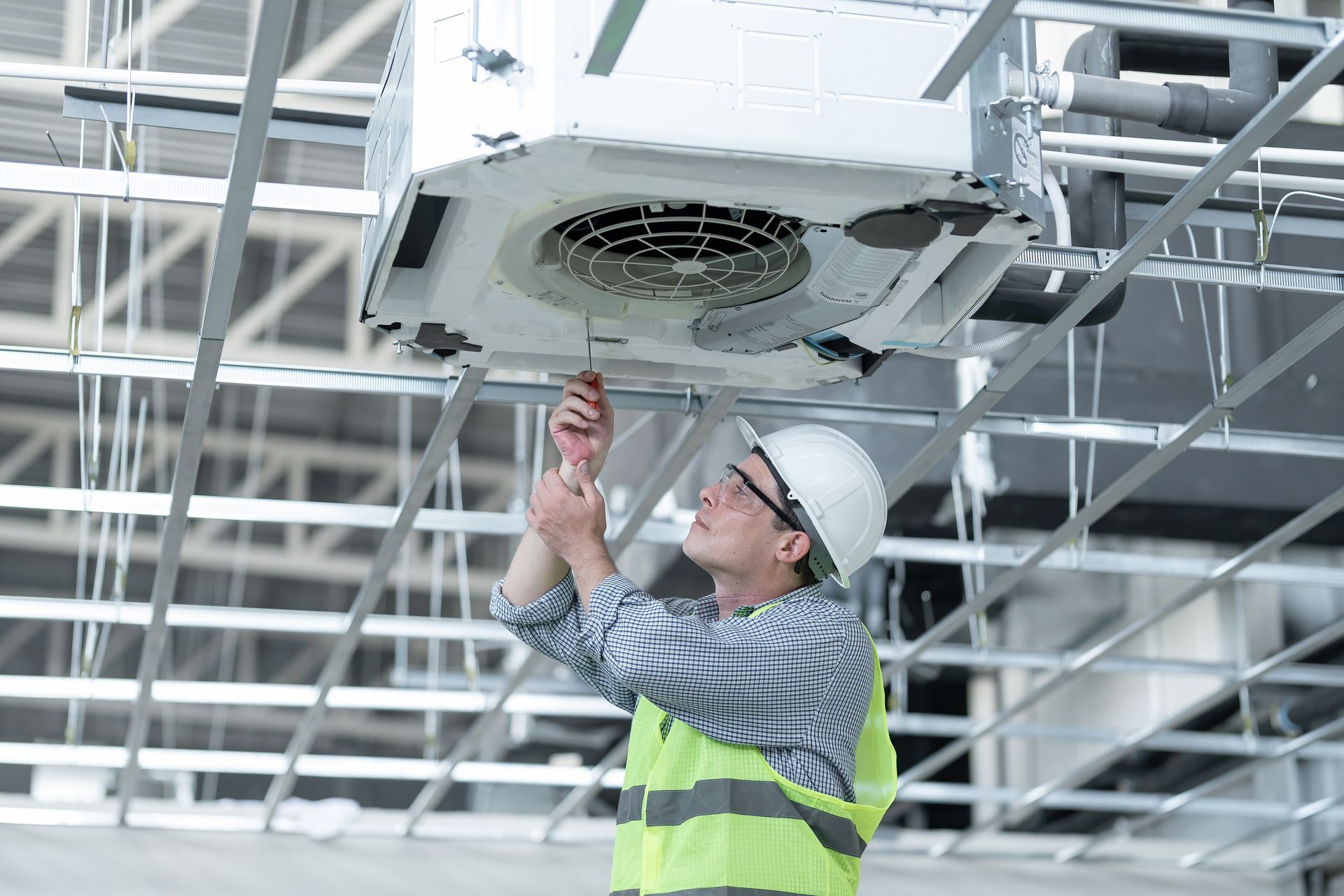 Worker in hard hat repairing a ceiling-mounted HVAC unit in a construction site Worker in hard hat repairing a ceiling-mounted HVAC unit in a construction site