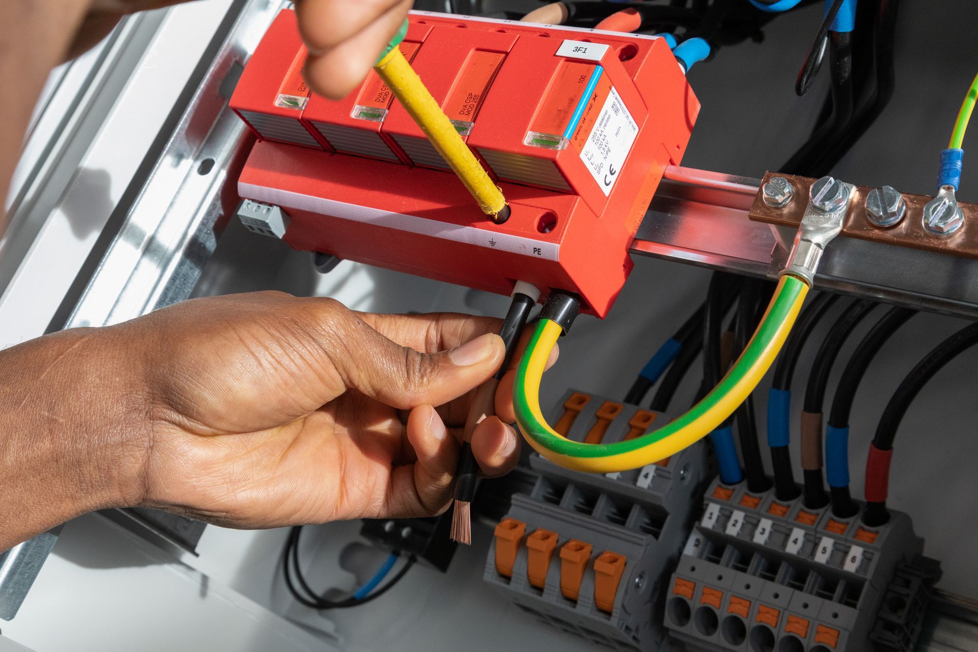 Hands wiring a yellow-green grounding cable in an electrical panel with a red tool and terminal blocks.