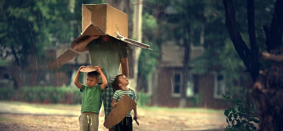 A man and two young boys holding cardboard boxes over their heads for shelter in a heavy downpour, with text overlay: How Weather in East Longmeadow MA Can Affect Your Storage Unit and How to Protect Your Belongings.