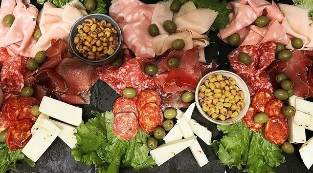 Un tagliere di salumi con vari tipi di carne, formaggi a fette, olive verdi e ciotoline di mais su un piatto scuro.