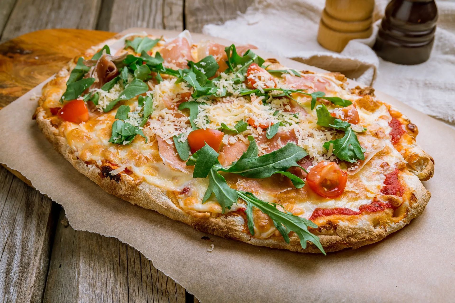 Pizza su pane piatto condita con formaggio fuso, prosciutto crudo, pomodorini e rucola fresca, servita su carta da forno.
