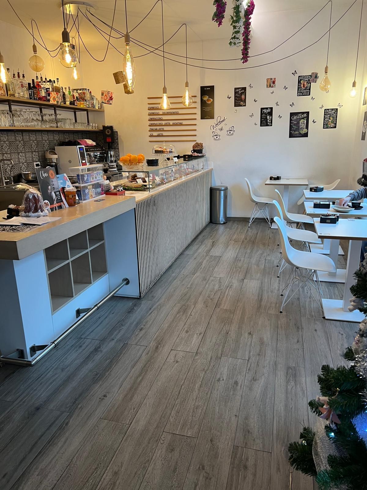 Un caffè luminoso con un lungo bancone, moderne sedie bianche e tavoli disposti lungo una parete con decorazioni appese.