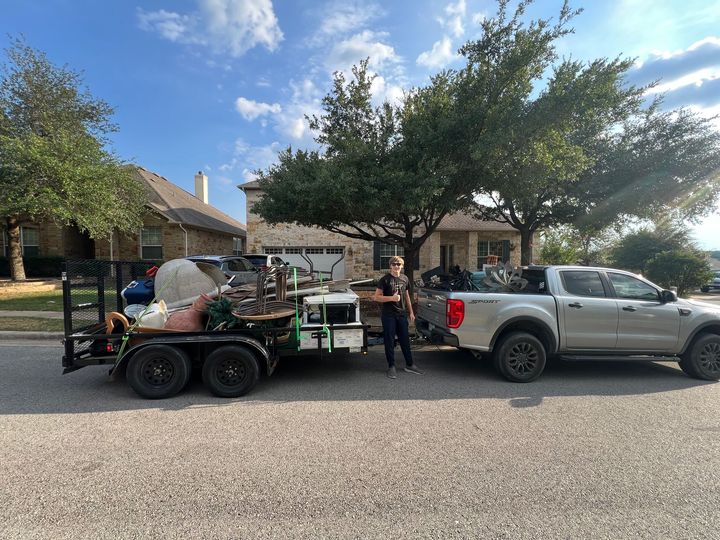 A person stands next to a truck pulling a utility trailer loaded with scrap metal and various items in a residential area.