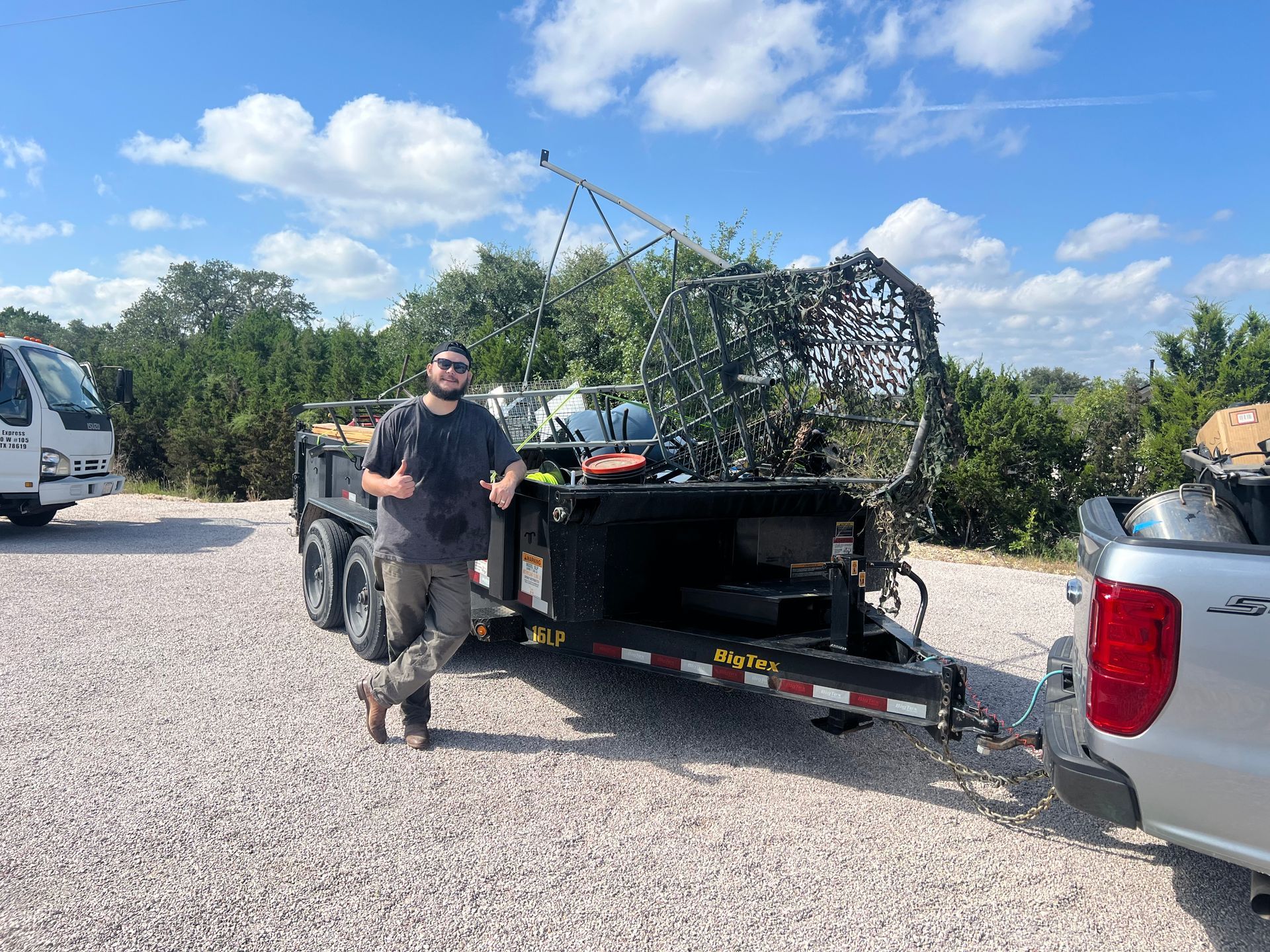 A person stands next to a trailer filled with equipment, including camouflage netting, on a gravel lot under a sunny sky.