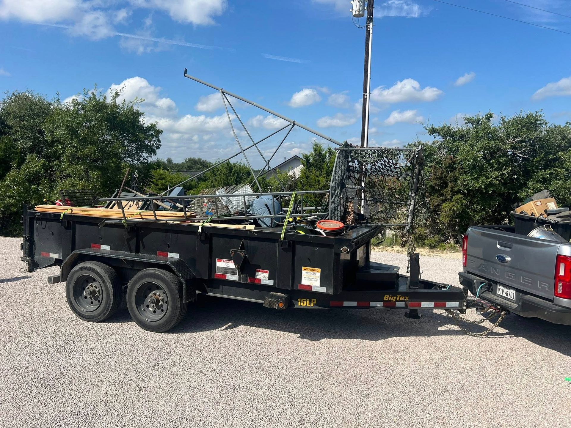 A black dump trailer loaded with construction materials and netting, hitched to a gray pickup truck on a gravel lot.