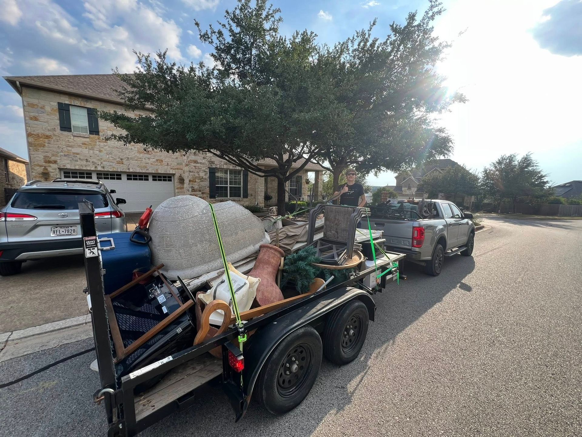 A trailer loaded with various items attached to a pickup truck parked on a residential street in front of a stone house.