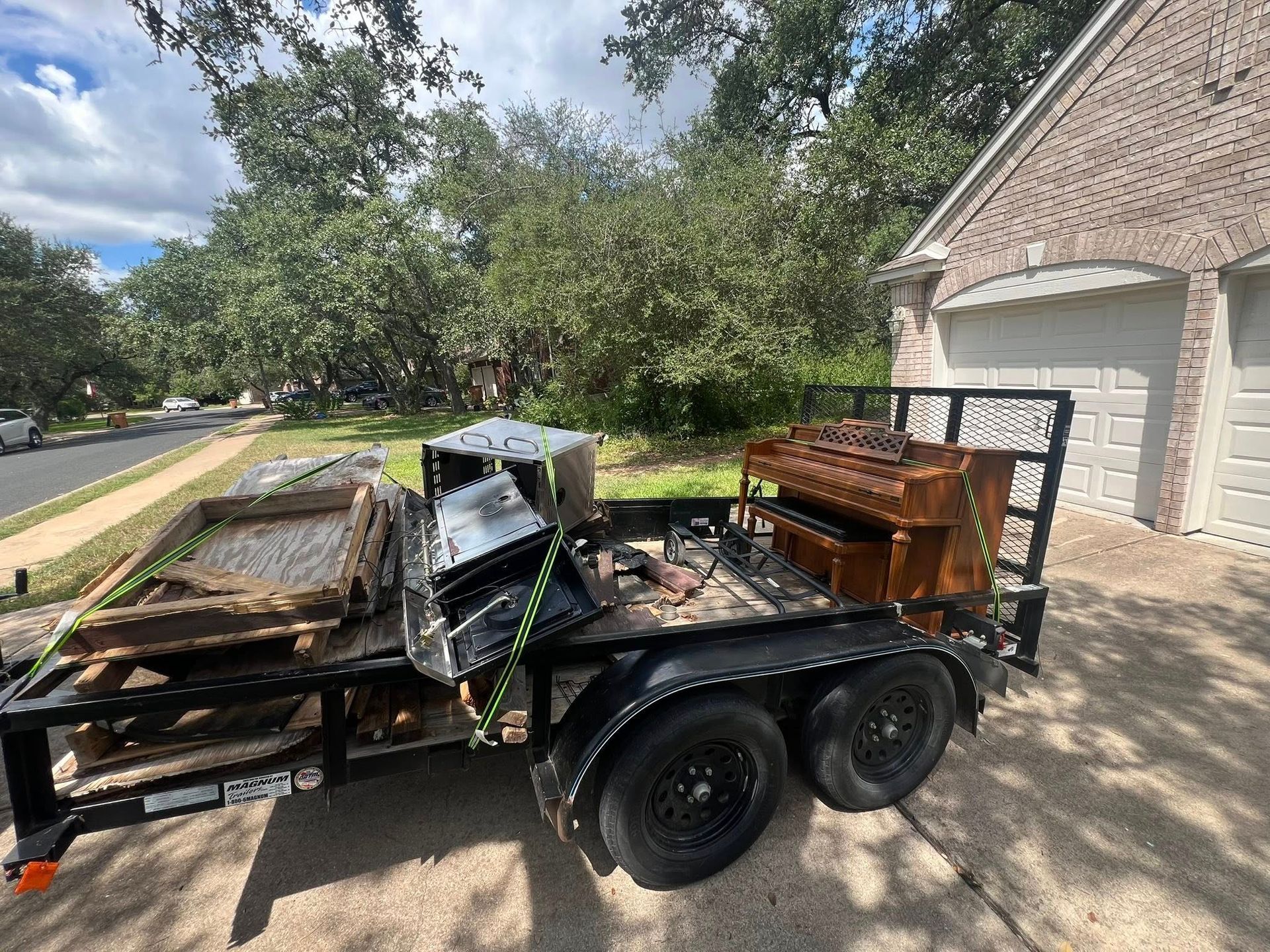 A utility trailer loaded with a wooden upright piano and scrap metal, parked in a residential driveway.