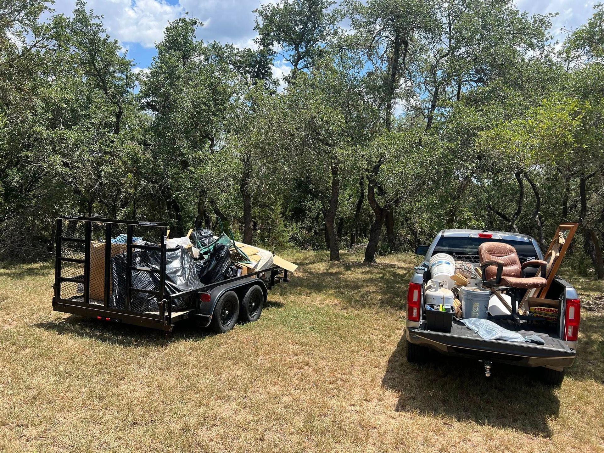 A flatbed utility trailer and a pickup truck loaded with assorted household items and debris parked in a grassy field.
