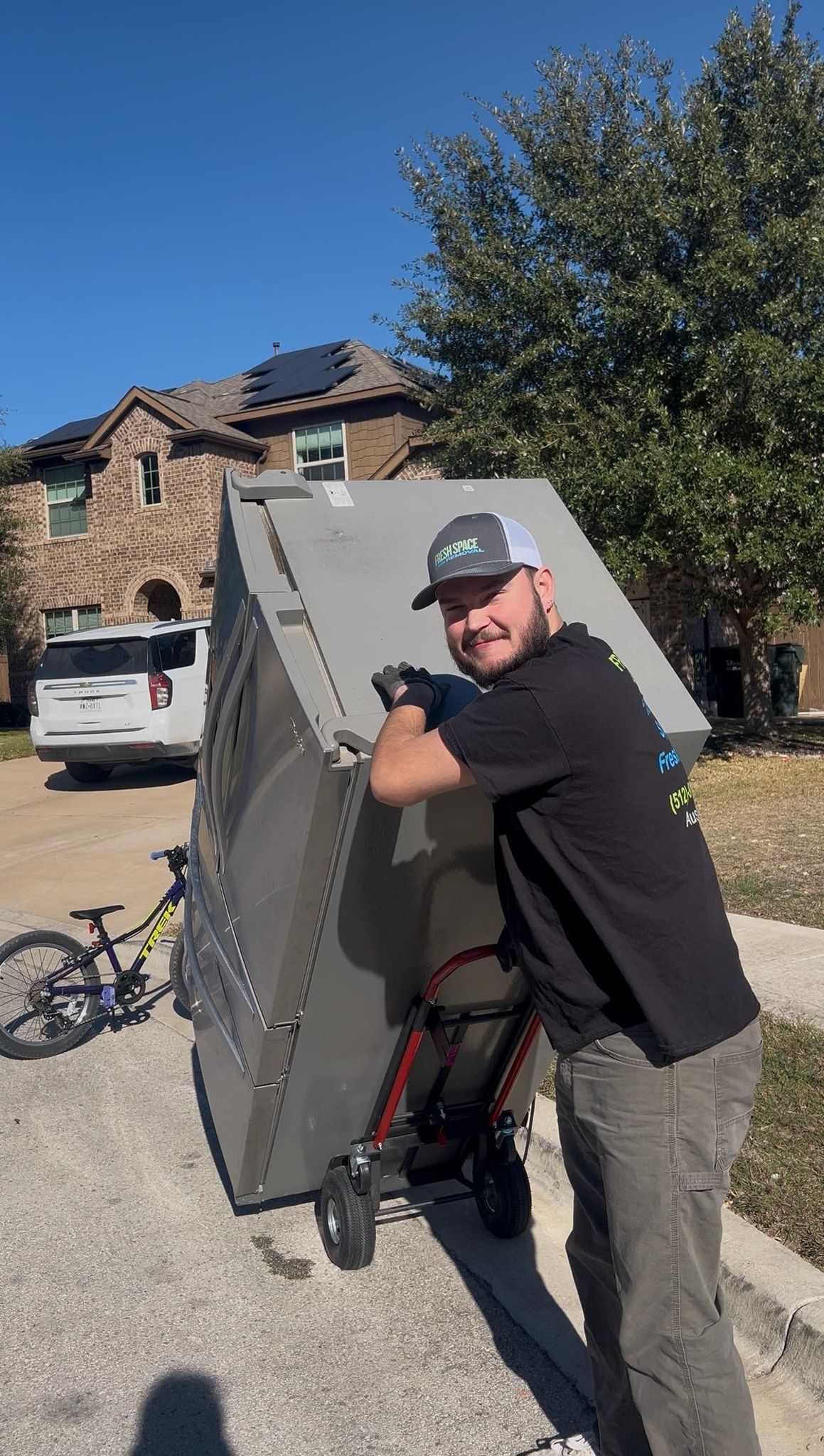 A person in a cap and black t-shirt pushes a large, grey, rectangular crate on a hand truck up a residential driveway.