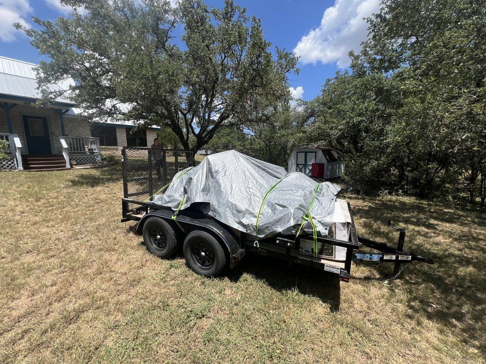 A black utility trailer carrying a large, gray, tarp-covered object parked on a grassy lawn in front of a house.