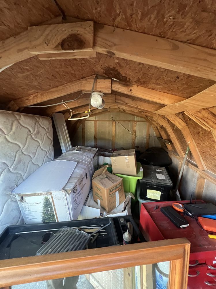 A view inside a cluttered wooden shed filled with stacked boxes, a mattress, a red tool chest, and miscellaneous items.