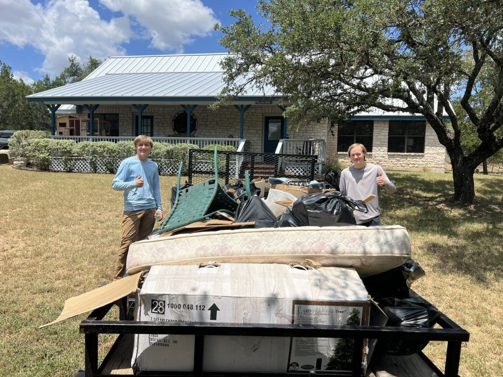 Two people stand next to a utility trailer packed with furniture, cardboard boxes, and bags in front of a stone house.