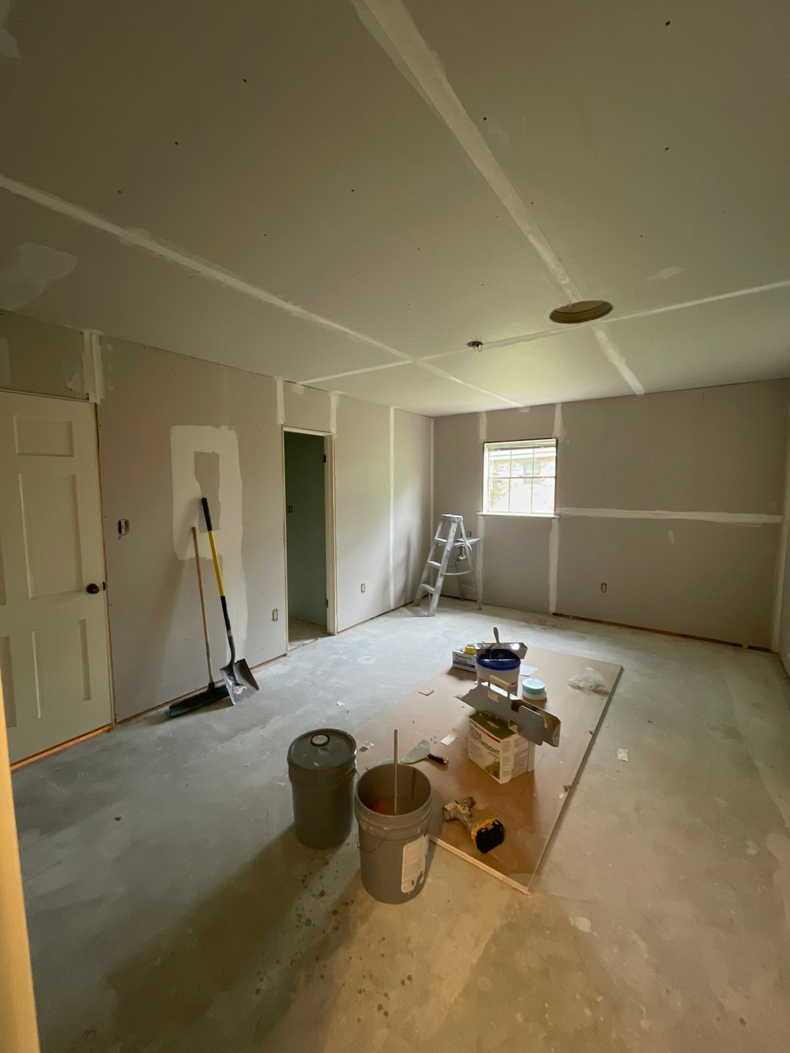 Room under construction, drywall installed on walls and ceiling. Buckets, tools, and materials scattered on the floor.