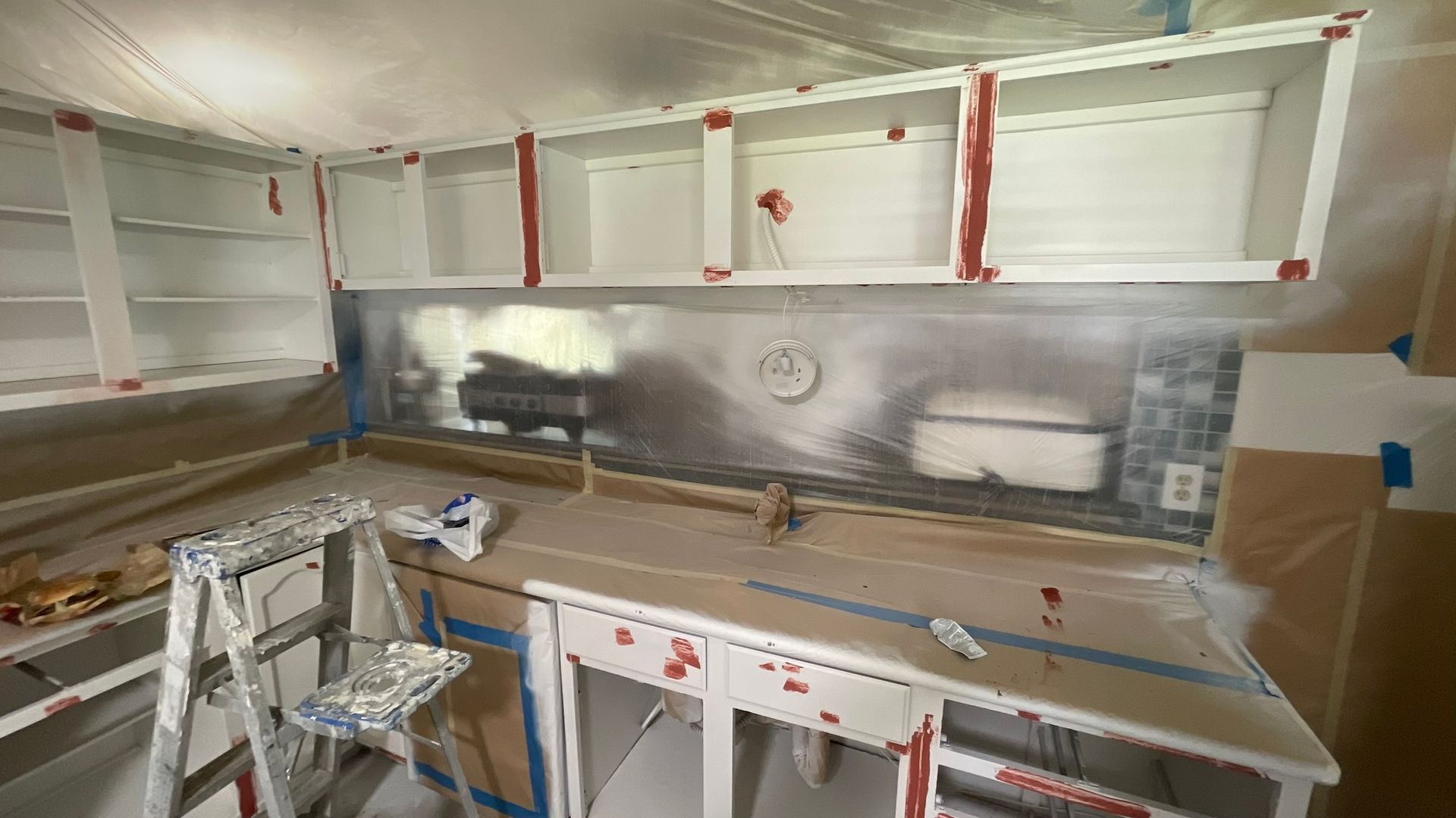 Kitchen cabinets being painted white; surfaces and appliances covered for protection.
