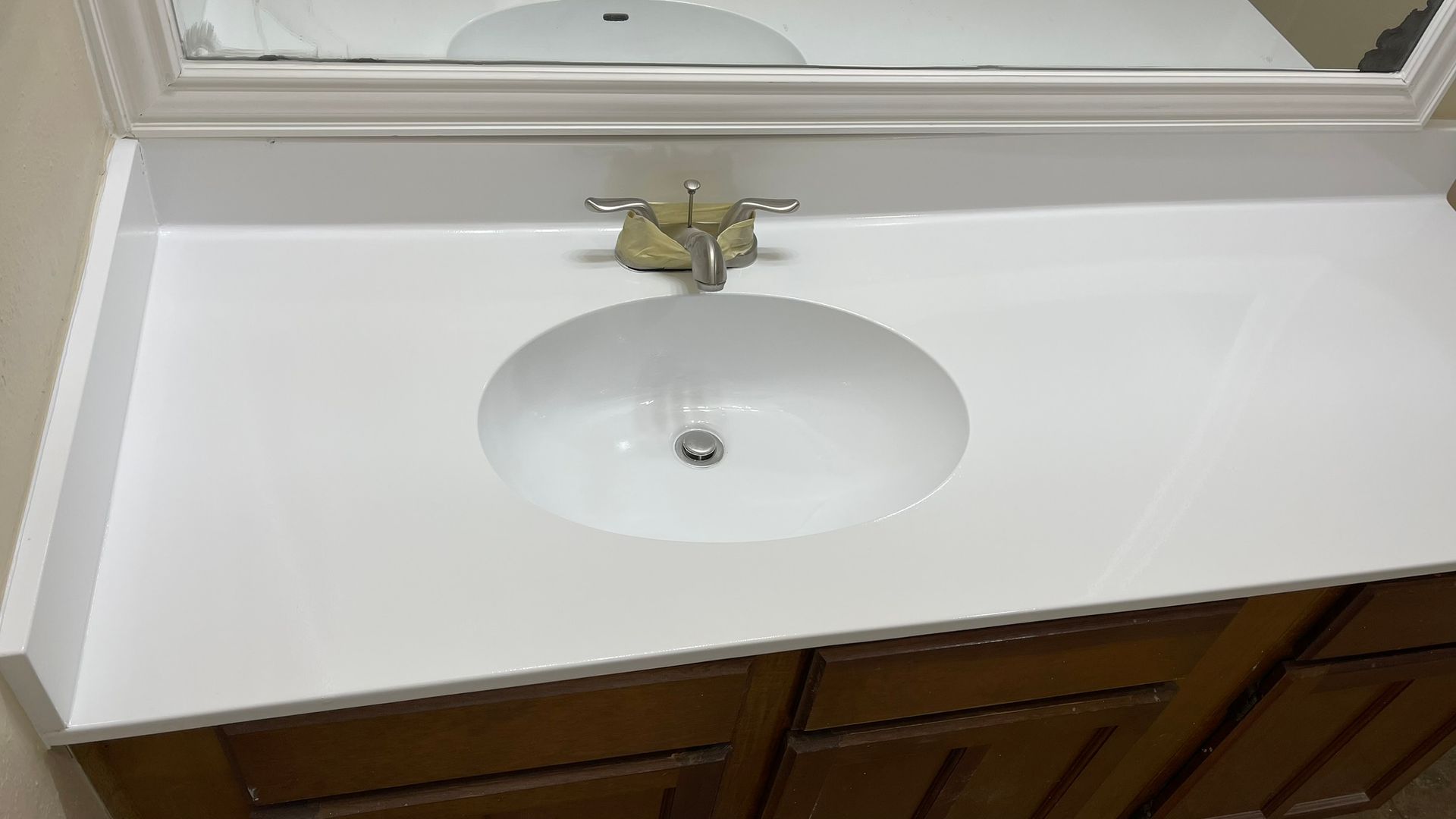 Bathroom vanity with white countertop and oval sink. Brown cabinet below with a mirror above.