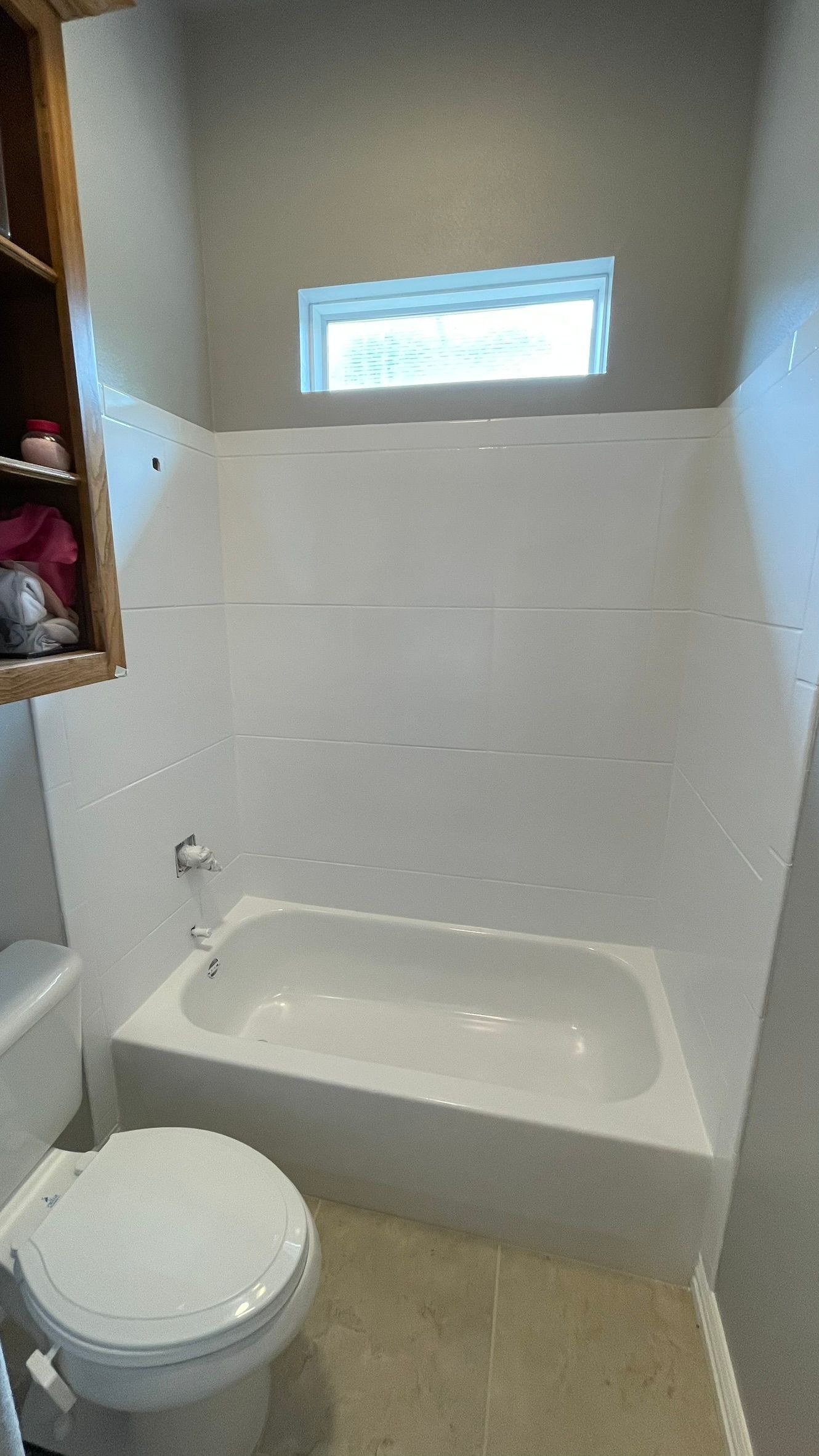 White bathtub and toilet in a bathroom with a window, tile floor, and a wooden cabinet.