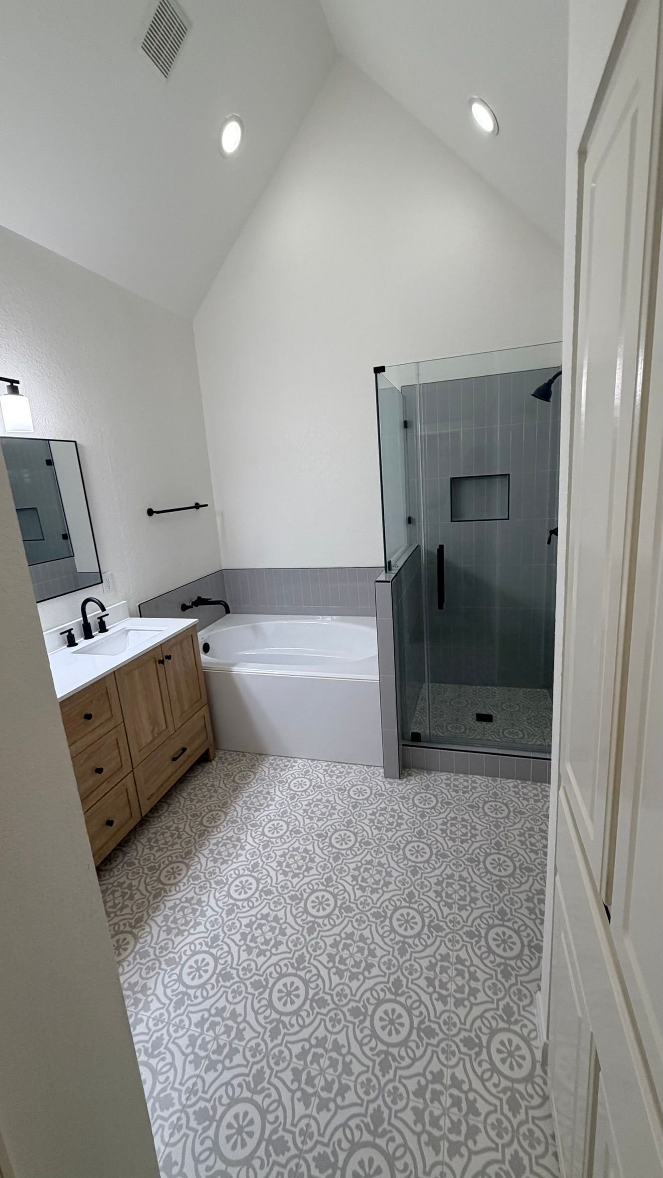Bright bathroom with white vaulted ceiling, patterned floor, double vanity, bathtub, and glass shower stall
