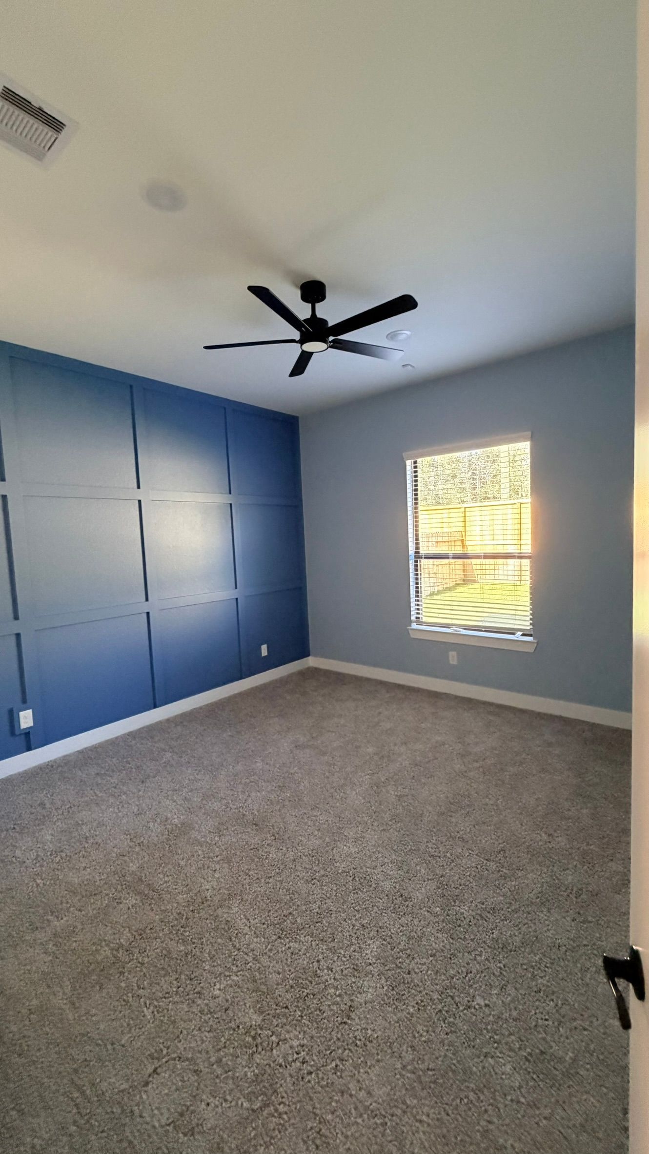Empty bedroom with blue accent wall, gray carpet, ceiling fan, and a window with blinds.