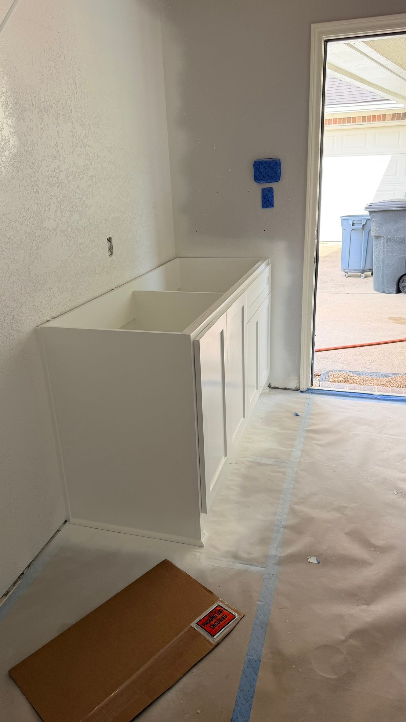 White cabinet along a plastered wall in an unfinished room, with open doorway and blue bins outside.