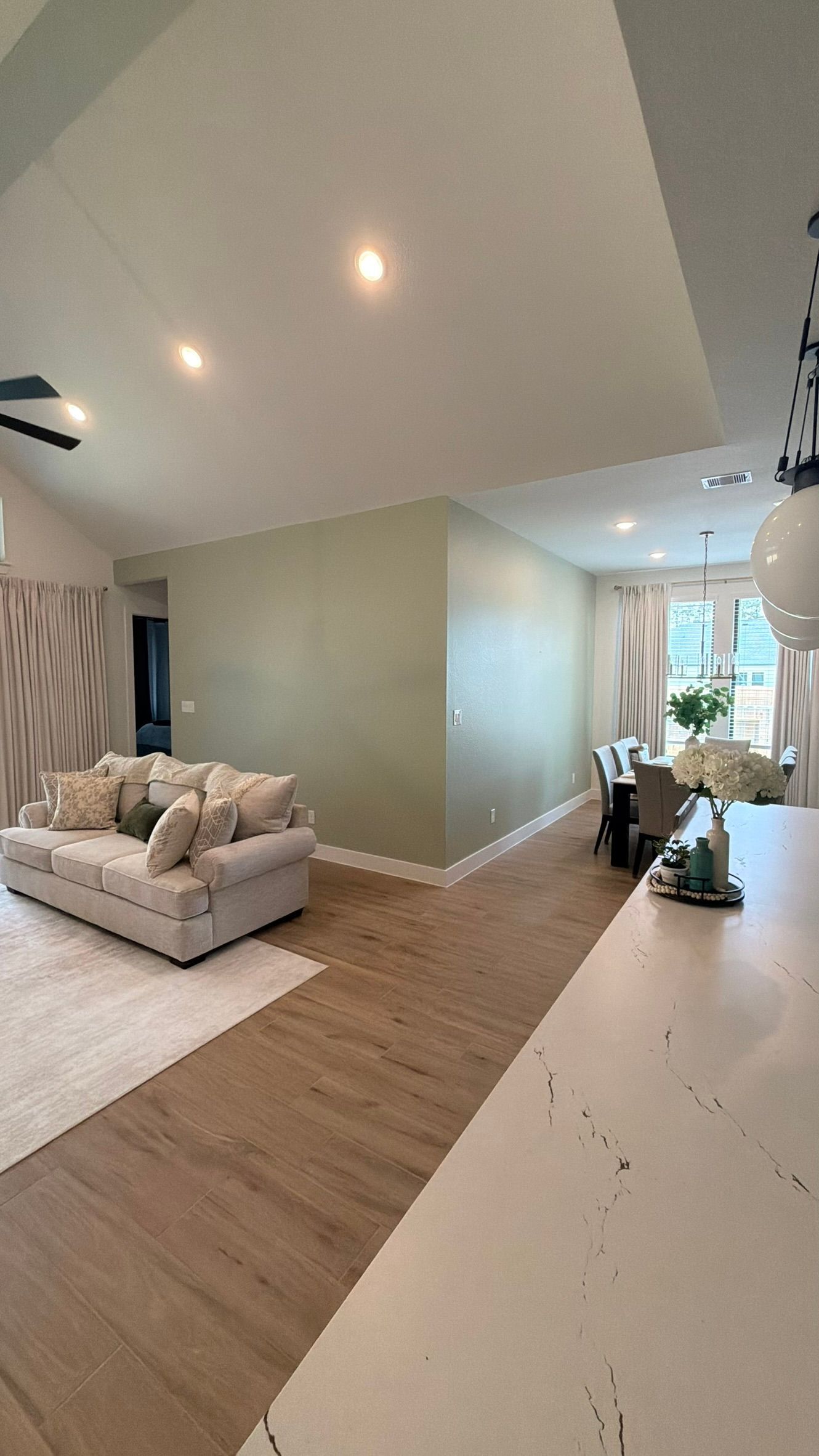 Bright living room with beige sofa, wood floors, recessed lights, and a large window near a kitchen counter.
