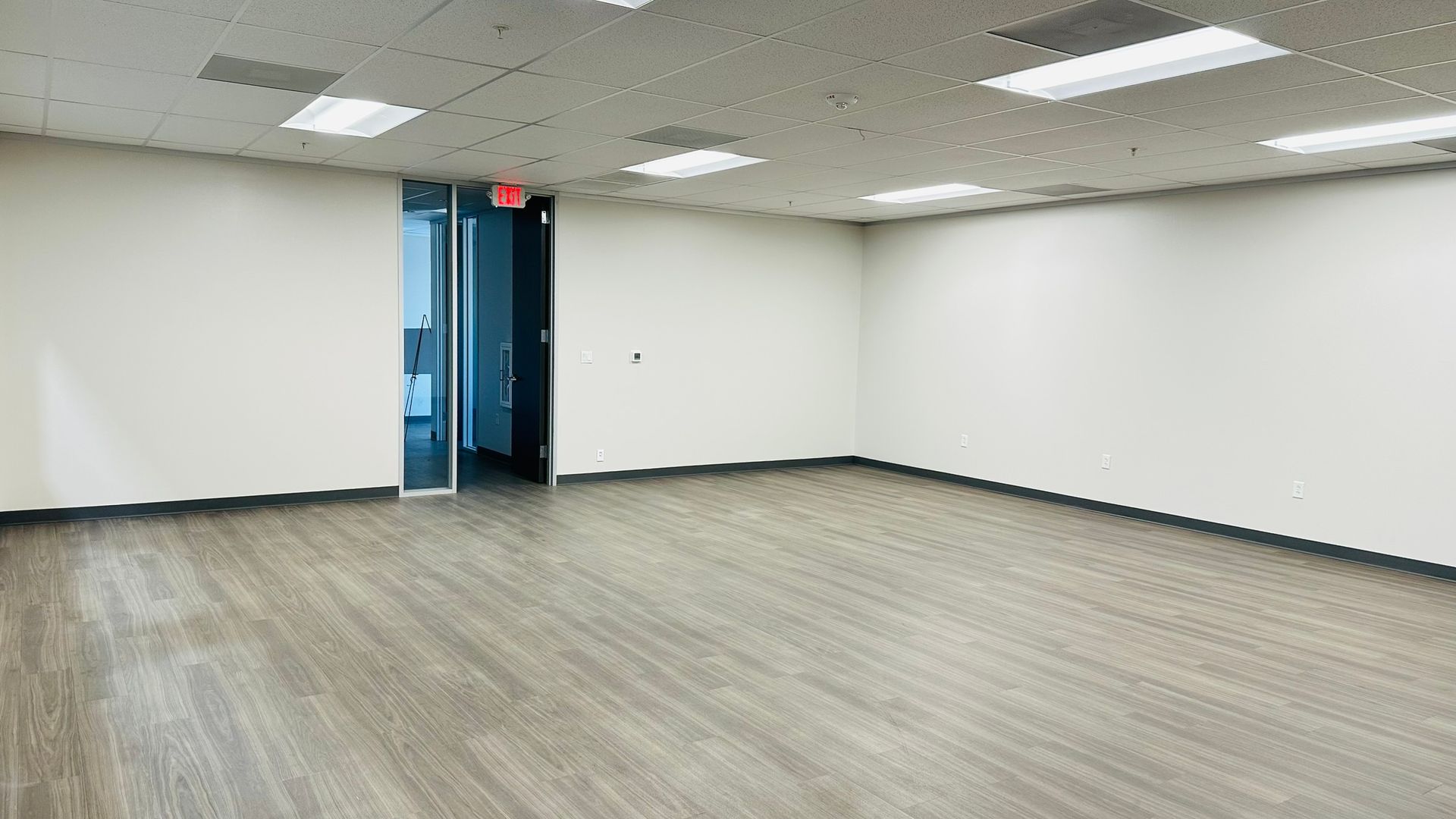Empty office room with gray wood-look flooring, white walls, and a door to another room.