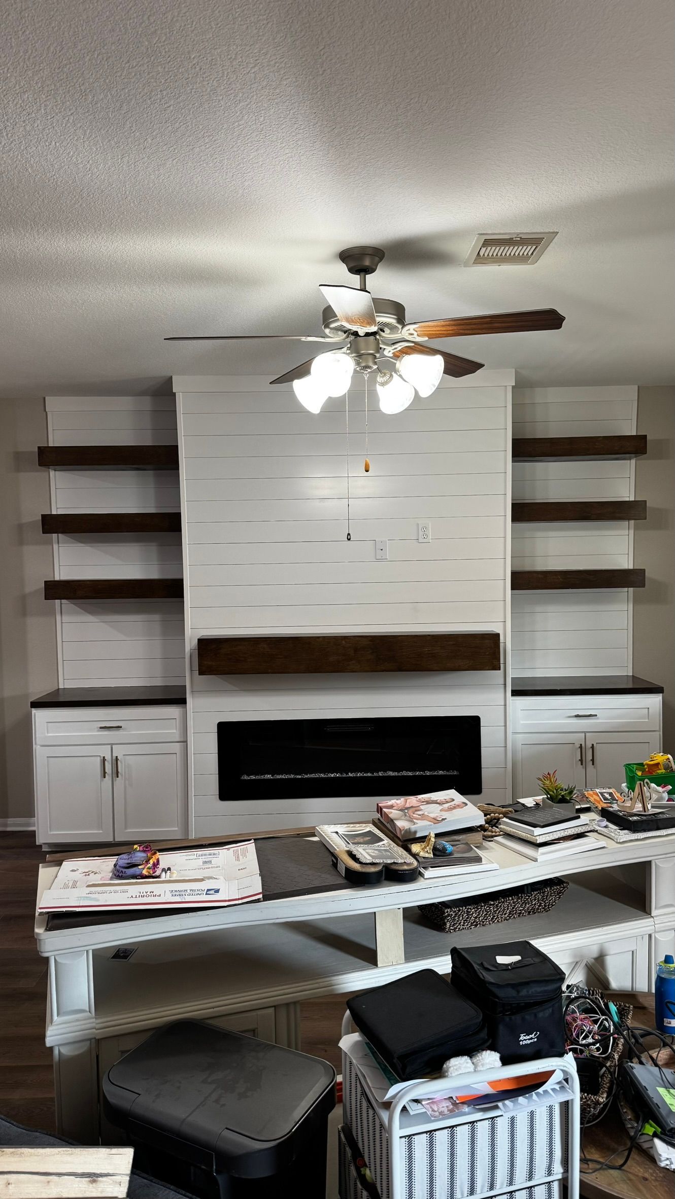 Living room with built-in shelves, fireplace, ceiling fan, and a cluttered coffee table and floor