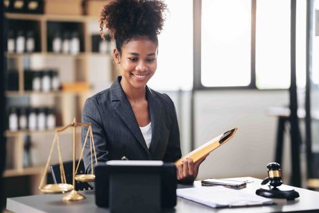 A woman is sitting at a desk holding a clipboard and a tablet.