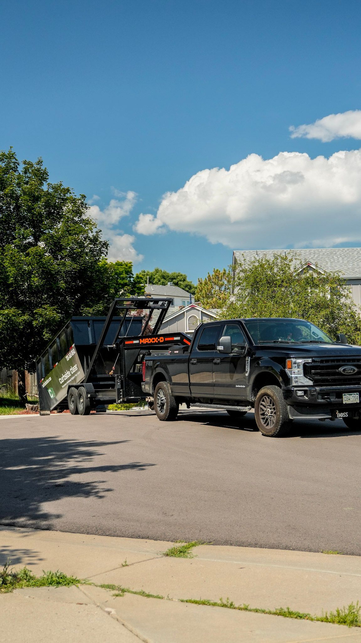 Black pickup and trailer parked on a suburban street under a blue sky with trees and clouds.