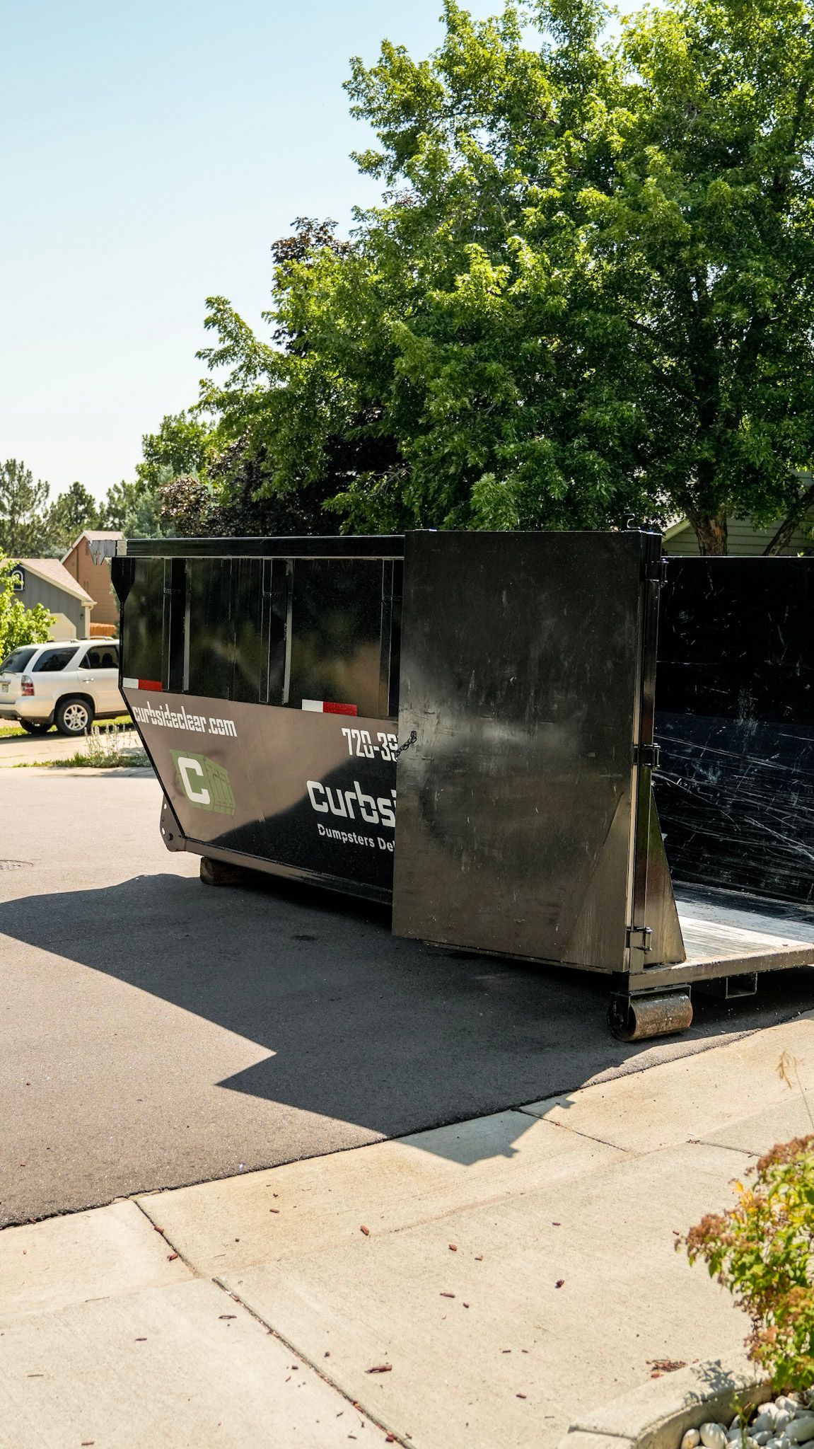 Large dark dumpster parked by a sidewalk under a leafy tree in a residential street.