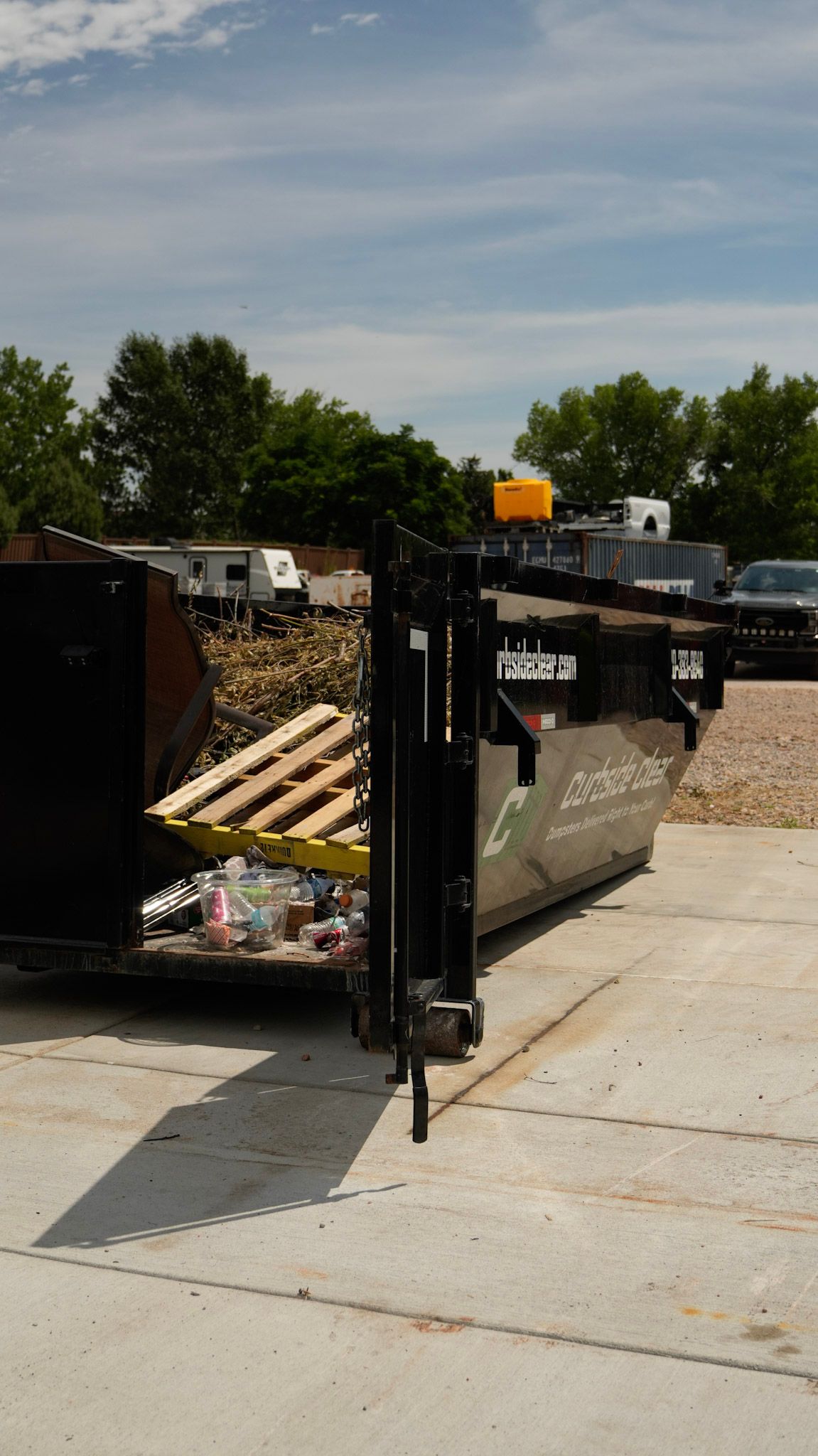 Open dumpster at a construction site, filled with wood and debris, beside a concrete path.