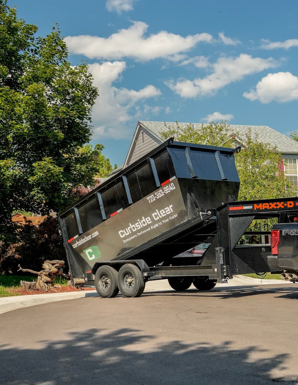 Large black dumpster truck tipping on a driveway under a blue sky, with trees and a house nearby