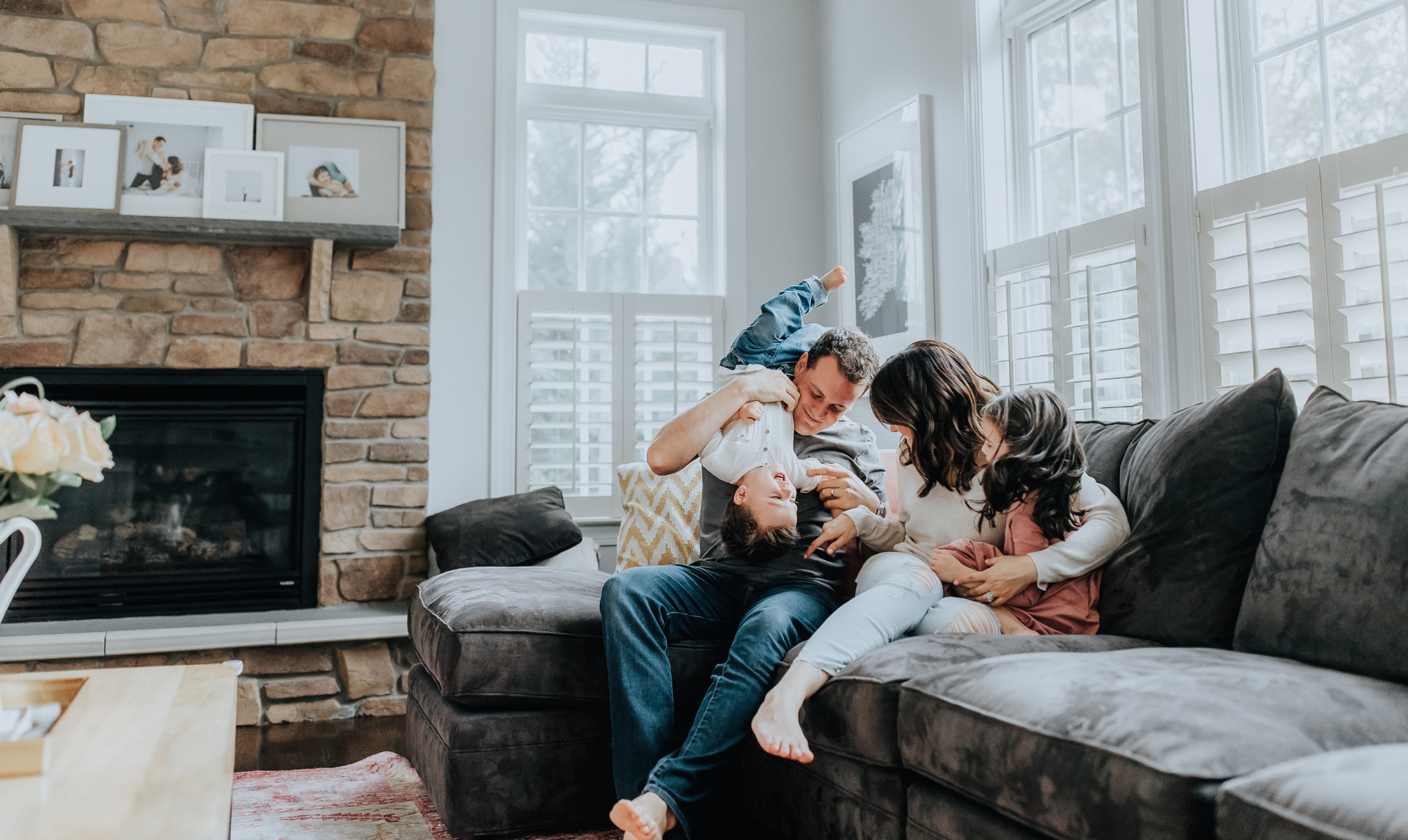 A family sits together on a gray sectional sofa in a living room with a stone fireplace, laughing and playing.