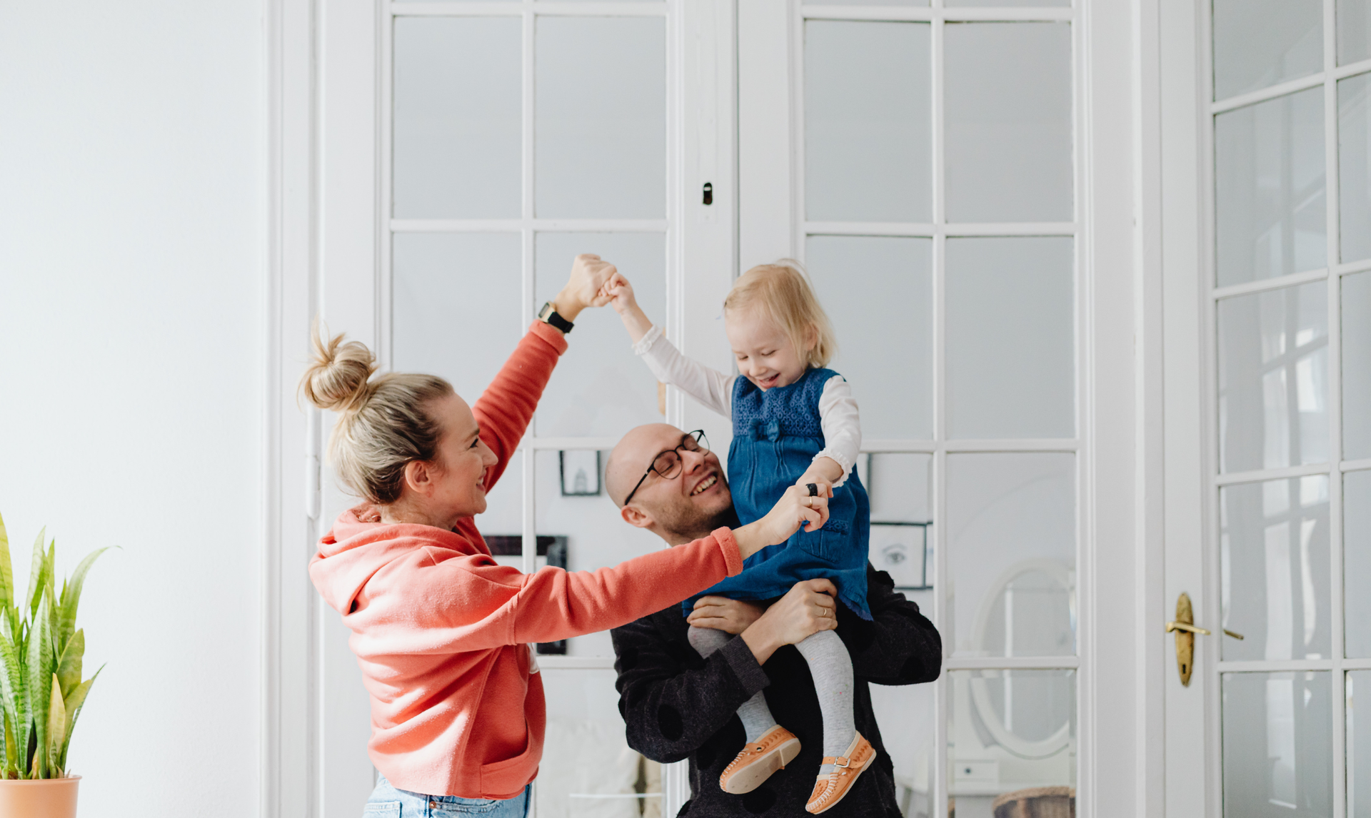 A person in a coral hoodie and a person in a black shirt lifting a child, all laughing in front of a glass-paneled door. A person in a coral hoodie and a person in a black shirt lifting a child, all laughing in front of a glass-paneled door.
