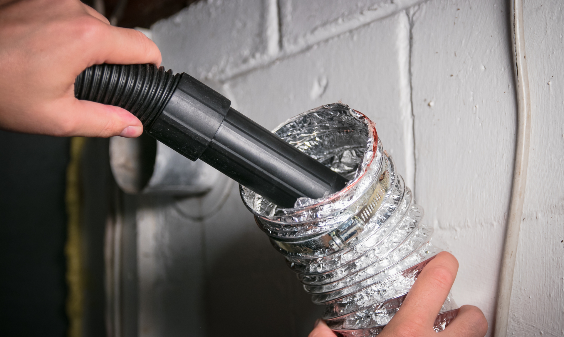 Hands use a vacuum attachment to clean the inside of a flexible silver dryer vent hose mounted on a white brick wall. Hands use a vacuum attachment to clean the inside of a flexible silver dryer vent hose mounted on a white brick wall.