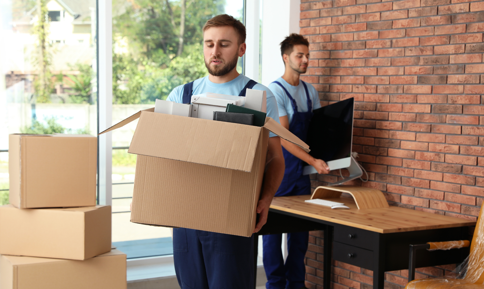 Two workers in blue uniforms move office supplies, one carrying a cardboard box while the other carries a computer monitor. Two workers in blue uniforms move office supplies, one carrying a cardboard box while the other carries a computer monitor.