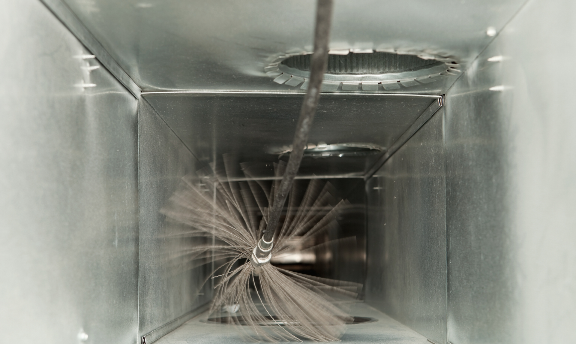 A circular bristle brush on a long rod inside a rectangular metal air duct during a cleaning process.