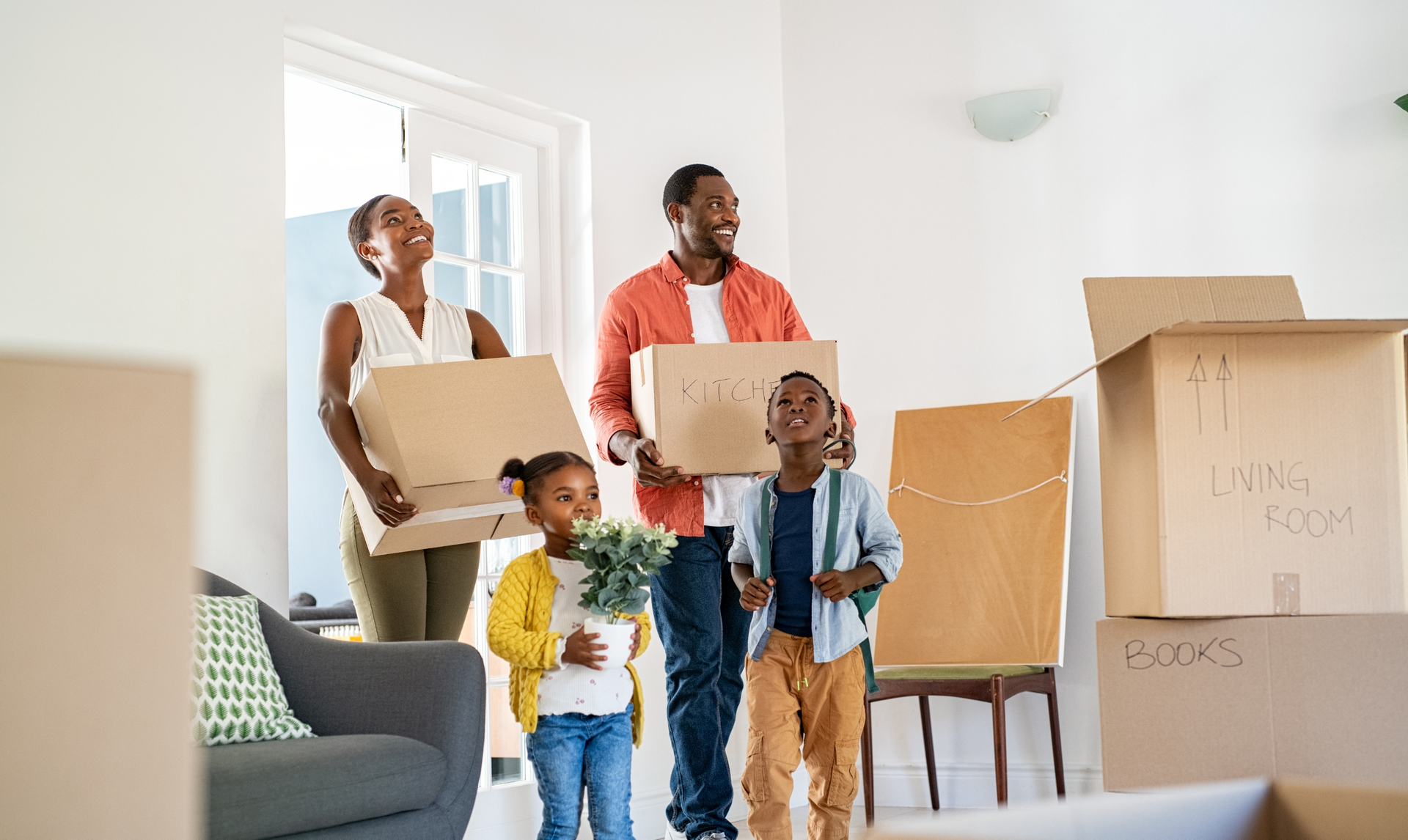A family stands in their new home holding moving boxes, looking up with smiles while surrounded by packing supplies. A family stands in their new home holding moving boxes, looking up with smiles while surrounded by packing supplies.