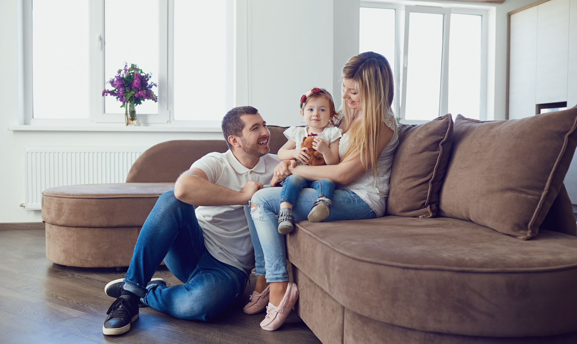 A small family sits together on a large brown sofa in a bright, modern living room. A small family sits together on a large brown sofa in a bright, modern living room.