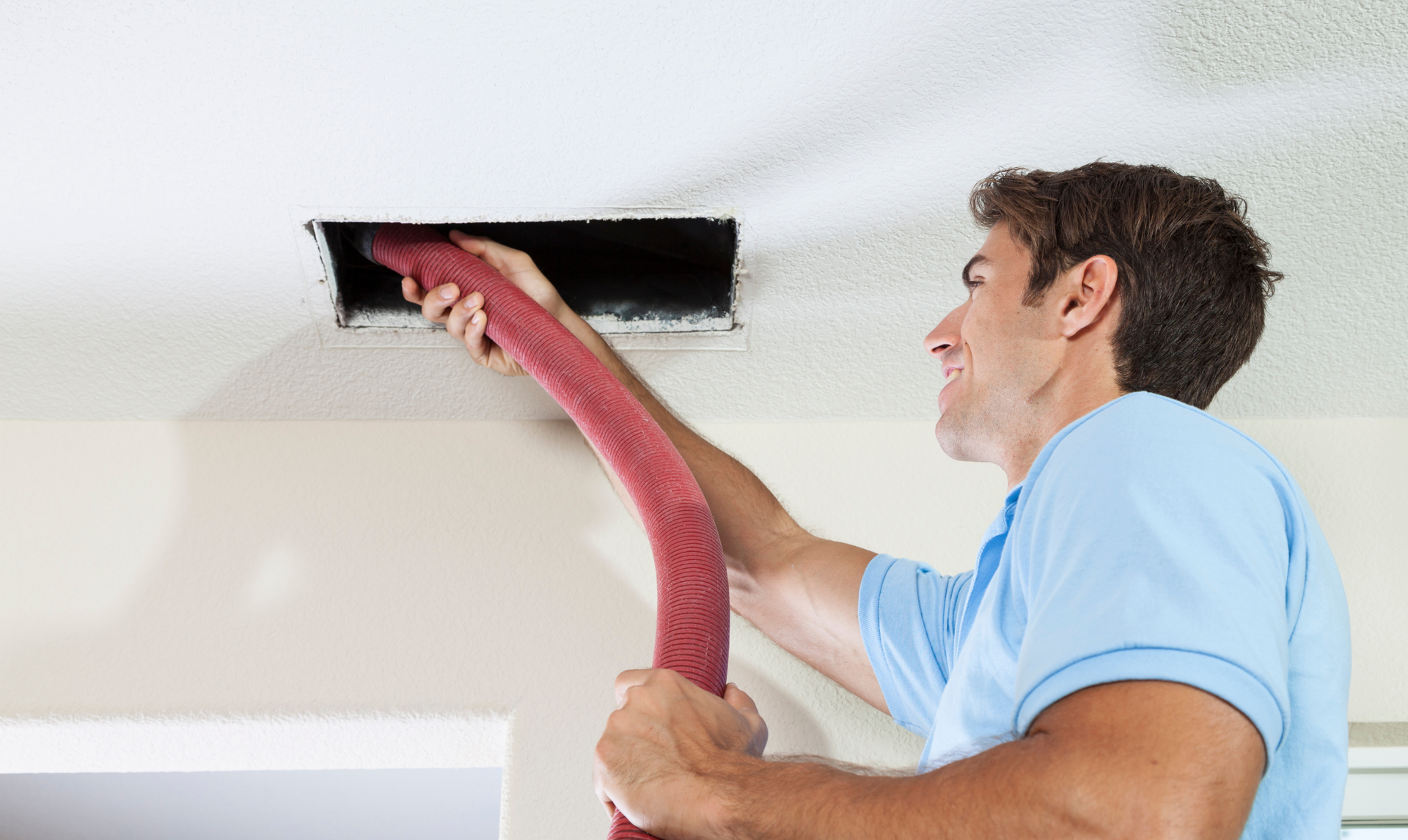 A person uses a vacuum hose to clean a rectangular air vent cut into a ceiling.