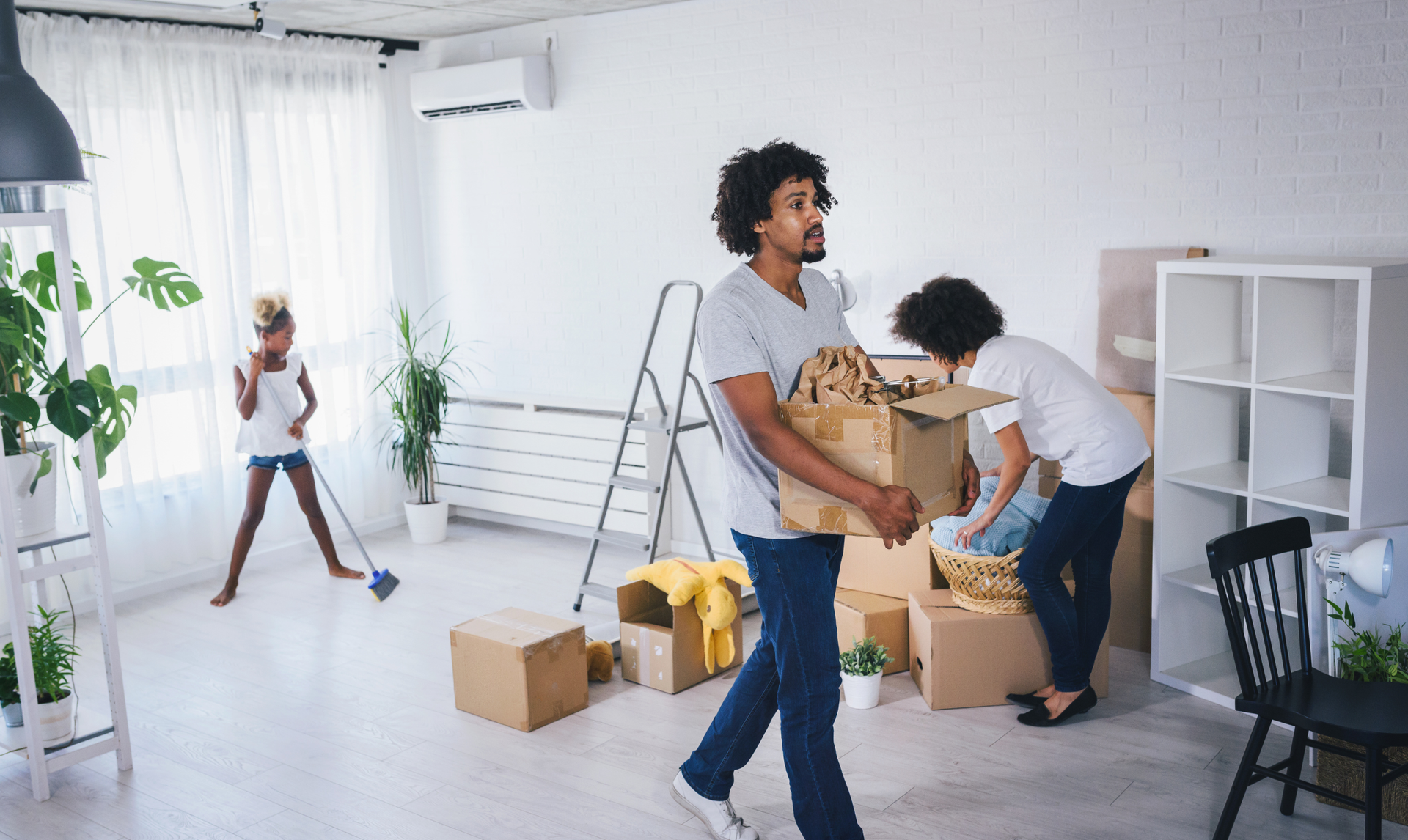 A family moving into a new house, with a person carrying a cardboard box while another sweeps the room.