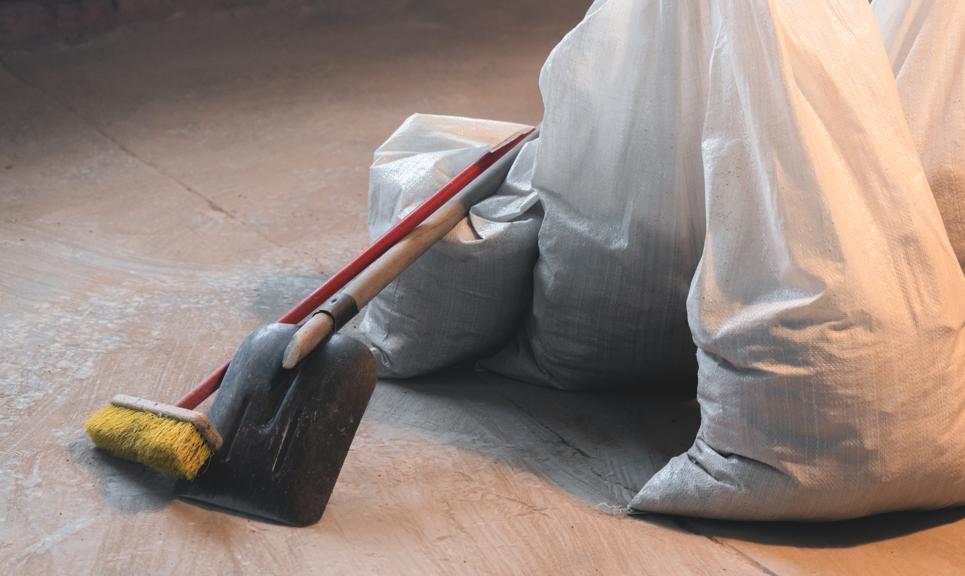 A yellow push broom and a black shovel lean against white sacks of building materials on a concrete floor.