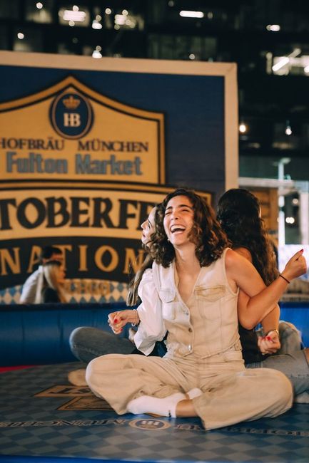 Woman sitting cross-legged on a stage, smiling in front of a Hofbräu München Oktoberfest sign.