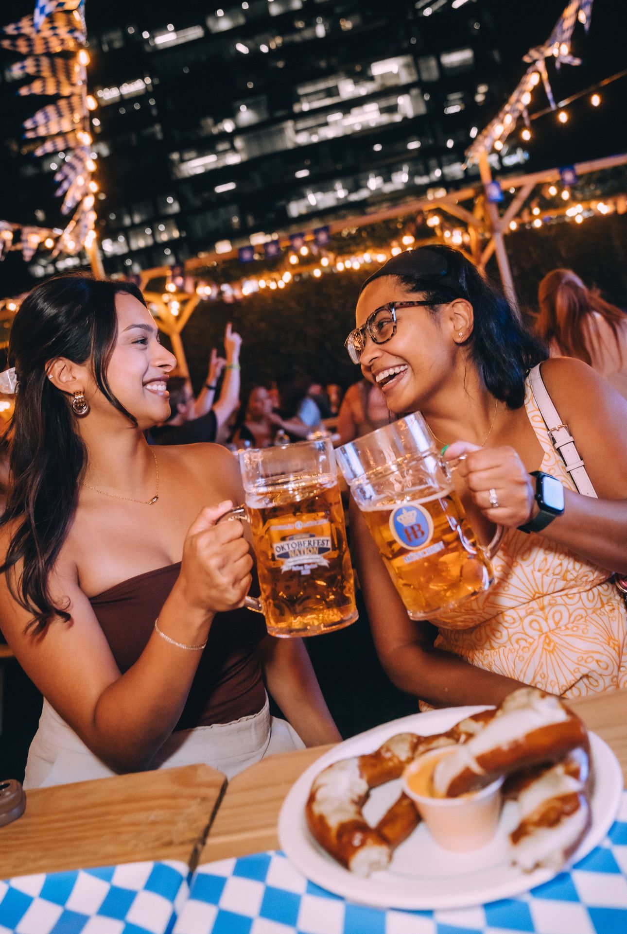 Two people toasting beer mugs at a nighttime outdoor table with plates of food and city lights behind them