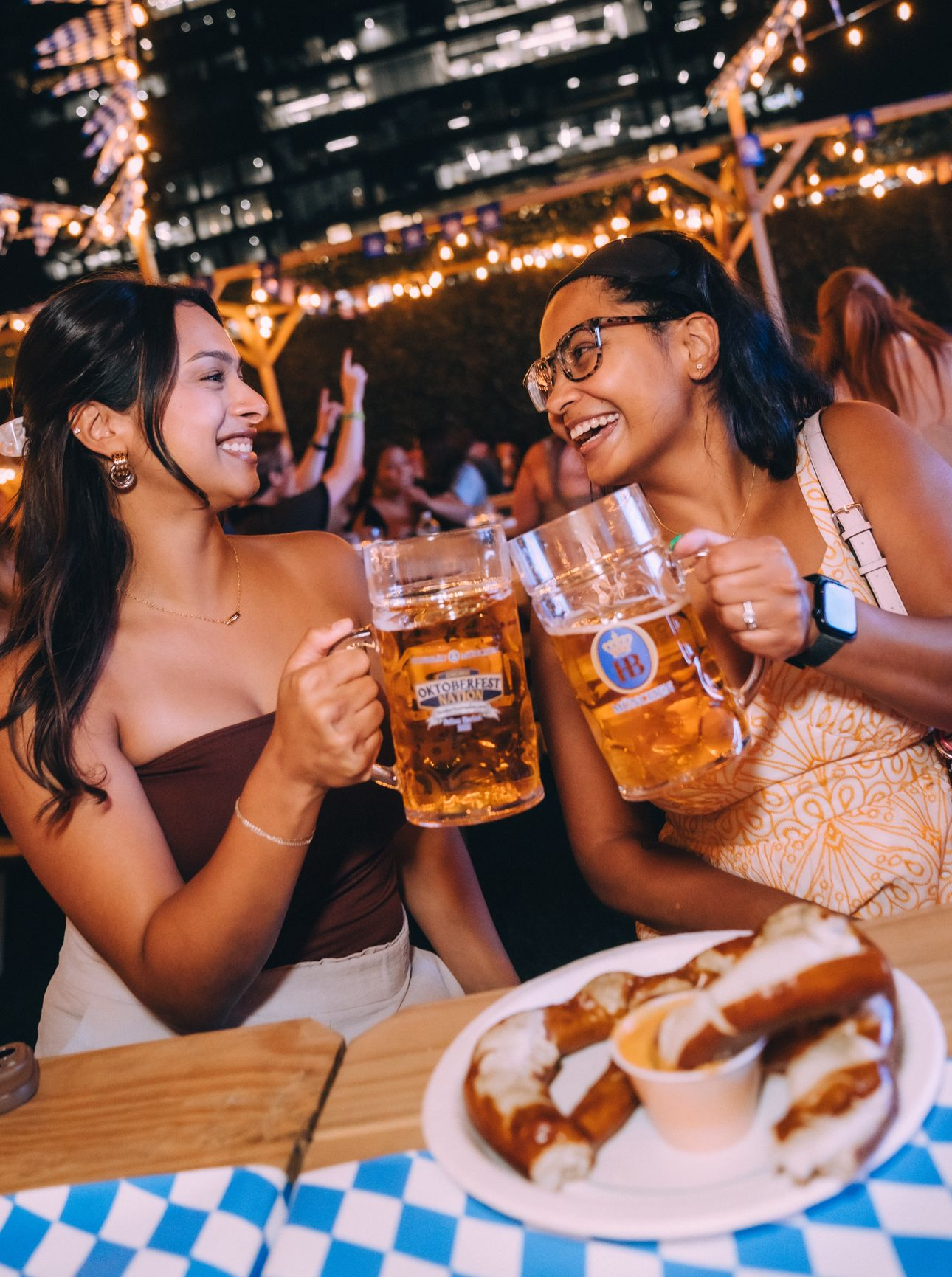 Two women clink beer mugs at a lively outdoor bar with plates of food in front of them.