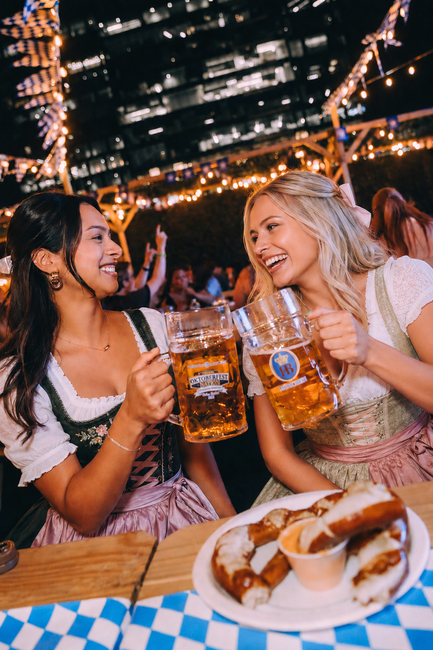 Two women clink beer mugs at a lively outdoor bar with plates of food in front of them.