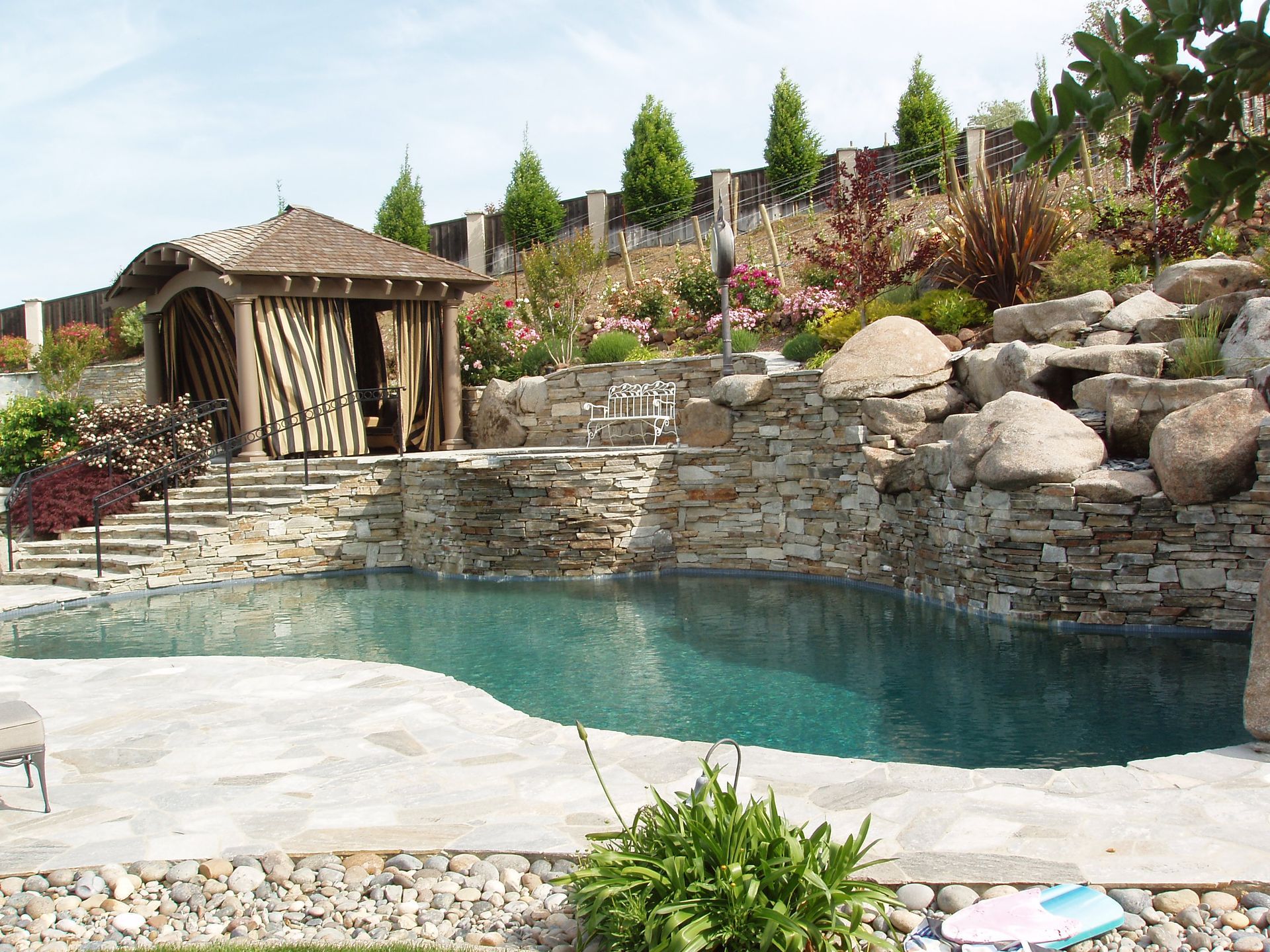 Backyard pool with turquoise water, stone waterfall, and a small wooden cabana amid landscaped rocks and flowers